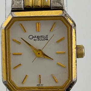 Puede incluir: Un reloj de pulsera Caravelle by Bulova con un bisel octogonal dorado y una correa plateada. La esfera del reloj es blanca con manecillas e indicadores dorados. El reloj tiene el texto "Caravelle by Bulova" y el número "40E32" en la esfera.
