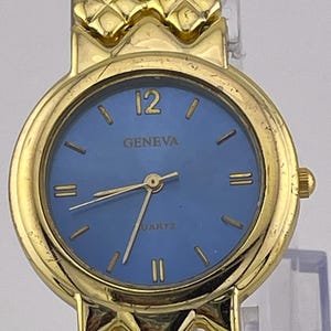 Puede incluir: Un reloj de cuarzo Geneva dorado con esfera azul. El reloj presenta manecillas e indicadores de hora dorados, con el número 12 en la parte superior. La correa del reloj tiene un diseño texturizado. La palabra "GENEVA" está impresa en la esfera.