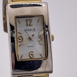 Puede incluir: Un reloj de pulsera rectangular con caja de metal plateado y esfera blanca. La esfera presenta números dorados en las posiciones 12, 3, 6 y 9, con el texto STUDIO TIME y QUARTZ impreso. La correa consta de eslabones plateados y de color crema.
