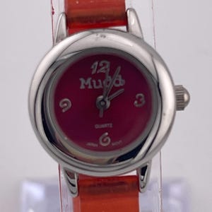 Könnte beinhalten: Eine silberfarbene Mudd-Quarzuhr mit rotem Zifferblatt und orangefarbenem Lederarmband. Das Zifferblatt zeigt den Markennamen und die Zahlen 12, 3, 6 und 9. Die Uhr hat eine kleine Krone und einen Schnallenverschluss.