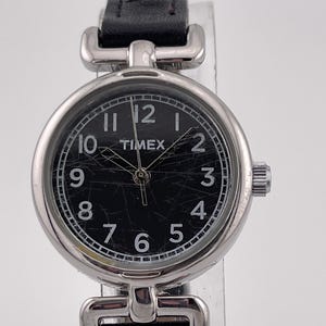 May include: A Timex wristwatch with a black face and silver-tone case. The watch has white numerals and hands, and a black leather band. The watch face is approximately 1 inch in diameter.
