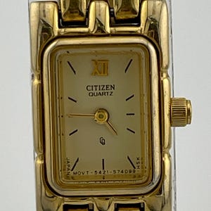 May include: Gold-toned Citizen Quartz watch with a rectangular face. The watch face is cream-colored with black hour markers and hands. The watch band is also gold-toned. The watch face displays the text "Citizen Quartz" and "Japan".