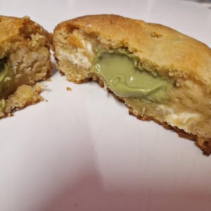 May include: A golden-brown baked biscuit split into two halves, revealing a soft, light green matcha-coloured cream filling and small white chunks of white chocolate embedded within the doughy, textured interior of the baked treat.