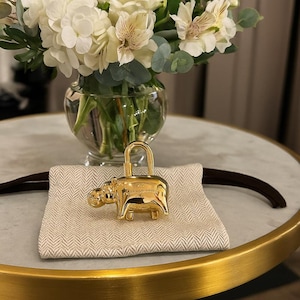 Puede incluir: Un candado dorado con forma de hipopótamo y asa curva, sobre una tela beige. El candado tiene texto grabado. Un ramo de flores blancas en un jarrón de cristal está al fondo.