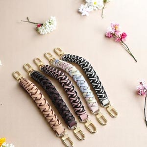 May include: Six braided purse handles in various colours: tan, black, dark brown, pink, light blue, and black and white. Each handle has gold-coloured clasps. The handles are arranged on a light beige surface with floral accents.