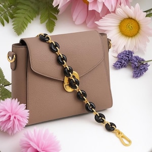 Könnte beinhalten: Eine taupefarbene Umhängetasche mit goldenem Verschluss und Beschlägen. Die Tasche hat eine dekorative Kette mit schwarzen, kreisförmigen Gliedern und goldenen Akzenten. Rosa Blumen und Grünzeug im Hintergrund.