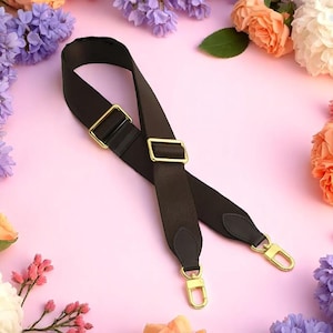 May include: A black and brown bag strap with gold-coloured metal hardware. The strap has adjustable buckles and swivel clasps for easy attachment. The background is a soft pink with floral accents.