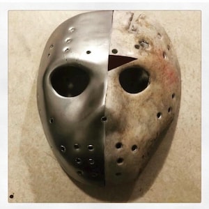 May include: A split-face mask, half metallic silver and half weathered beige, with dark eye holes and numerous small circular perforations. The mask is a prop or costume piece, likely for a horror theme.