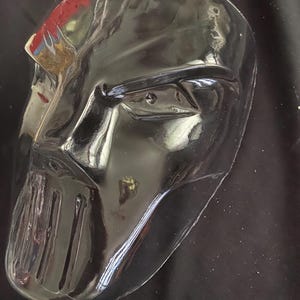 Puede incluir: Una máscara de plástico negro con un diseño de rostro detallado. La máscara tiene una barbilla texturizada en relieve y un solo agujero para el ojo.
