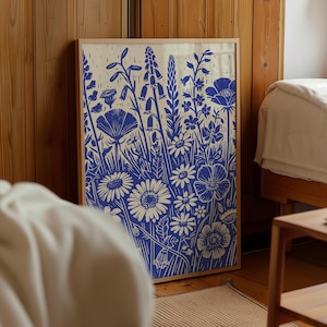 May include: A framed print of a blue and white floral design. The print features a variety of flowers, including daisies, poppies, and bellflowers. The flowers are arranged in a loose, organic pattern, and the background is a light cream color.