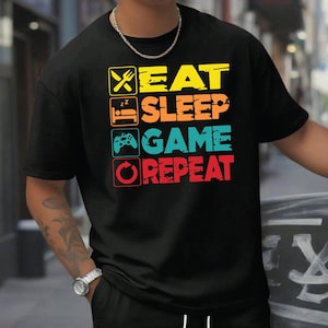 May include: Black t-shirt and shorts set. The t-shirt displays the text "EAT SLEEP GAME REPEAT" in yellow, orange, teal, and red. The set includes black shorts with a cargo pocket.