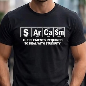May include: A black short-sleeved t-shirt featuring a graphic of four periodic table elements spelling S Ar Ca Sm. Below the graphic, the text reads: THE ELEMENTS REQUIRED TO DEAL WITH STUDIPITY. The design is printed in white ink on the front of the shirt.
