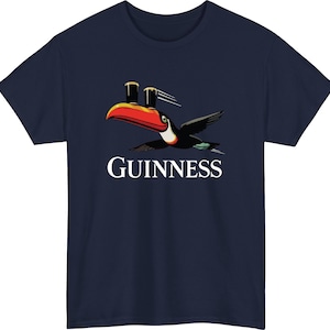 May include: Navy blue t-shirt featuring a toucan carrying two pints of stout, with the word "GUINNESS" in white below. The toucan has a red beak, black wings, and a white belly. A classic design for fans of the Irish stout.