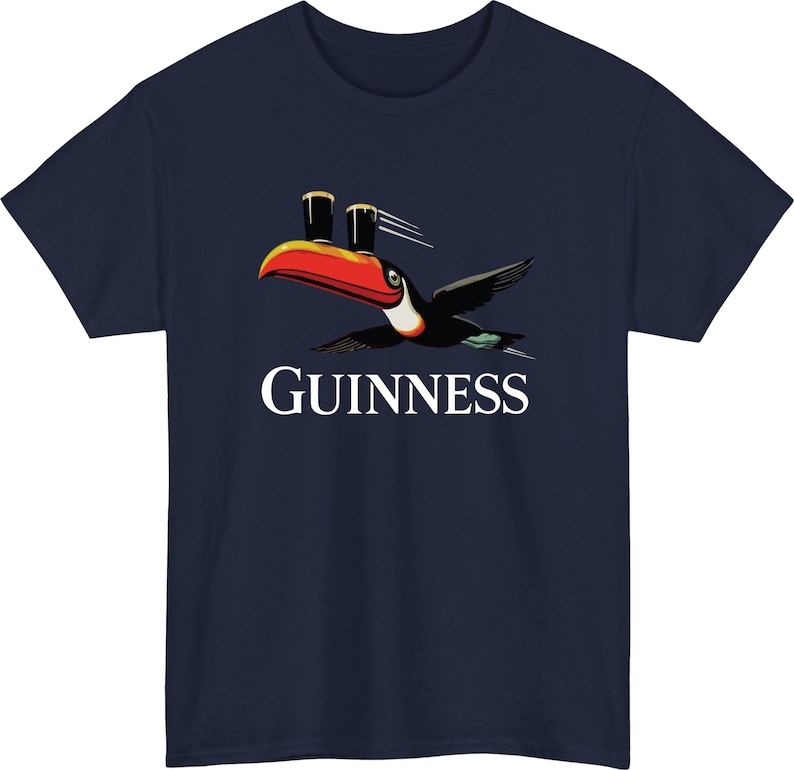 May include: Navy blue t-shirt featuring a toucan carrying two pints of stout, with the word "GUINNESS" in white below. The toucan has a red beak, black wings, and a white belly. A classic design for fans of the Irish stout.