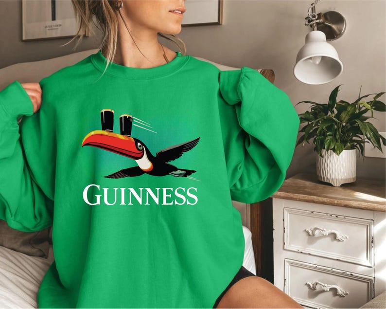 May include: Green sweatshirt featuring a toucan with a red and black beak, wings spread, and two pints of Guinness on its head. The word "GUINNESS" is printed in white below the bird. The sweatshirt is a solid green color.