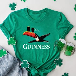 May include: A green t-shirt featuring a toucan carrying two pints of Guinness. The word "GUINNESS" is printed in white below the toucan. Shamrock cutouts and a pair of jeans are also in the image.