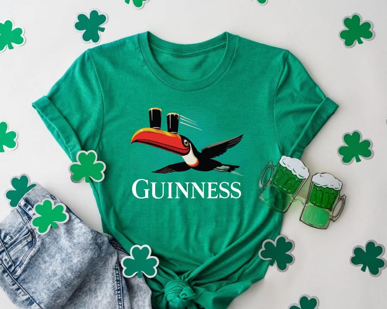 May include: A green t-shirt featuring a toucan carrying two pints of Guinness. The word "GUINNESS" is printed in white below the toucan. Shamrock cutouts and a pair of jeans are also in the image.