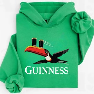 May include: Green hooded sweatshirt featuring a toucan with a red and orange beak, carrying two pints of Guinness. The word "GUINNESS" is printed in white below the bird. The sweatshirt has a drawstring hood and long sleeves.