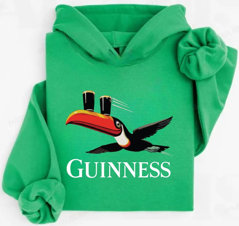 May include: Green hooded sweatshirt featuring a toucan with a red and orange beak, carrying two pints of Guinness. The word "GUINNESS" is printed in white below the bird. The sweatshirt has a drawstring hood and long sleeves.