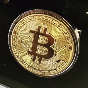 May include: A gold-colored metal coin with a raised image of a bitcoin symbol. The coin has a silver-colored rim and is surrounded by a black background.