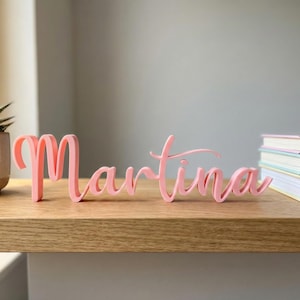 Può includere: Un nome rosa, in stile corsivo, "Martina", è esposto su una mensola di legno. Una piccola succulenta in un vaso beige e una pila di libri sono anche sulla mensola. Il nome è un oggetto decorativo.