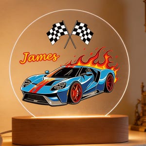 May include: A circular illuminated acrylic panel features a blue sports car design with red details and flames. The name "James" is above the car, and two checkered flags are at the top. The panel is set on a wooden base, creating a decorative night light.
