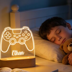 May include: A lit-up, clear acrylic controller-shaped night light with the name "Ethan" engraved on it, sitting on a wooden base. A child sleeps in bed with a teddy bear. A comic book is open on the nightstand.
