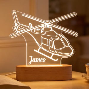 May include: A helicopter-shaped night light, illuminated and etched with the name "James". The light is made of clear acrylic and sits on a wooden base. A book and a teacup are on the table.
