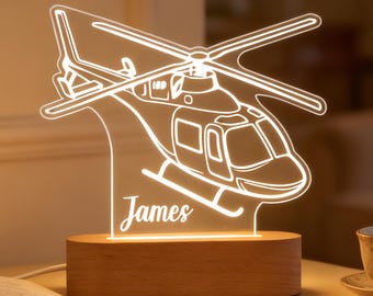 May include: A lit-up helicopter-shaped night light with the name "James" etched on the base. The light is made of clear acrylic and sits on a wooden base. A book and a teacup are on the table.