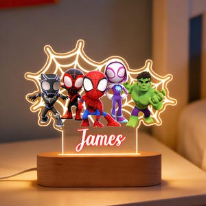 May include: A wooden night light with a glowing spiderweb design, featuring superhero figures. The light includes Black Panther, two Spider-Man characters, Spider-Gwen, and the Hulk. The name "James" is illuminated on the base.