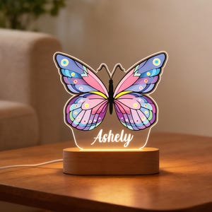 May include: A colorful butterfly-shaped night light with a wooden base. The butterfly has pink, purple, and blue wings with the name "Ashely" illuminated on the base. The lamp is on a wooden table.