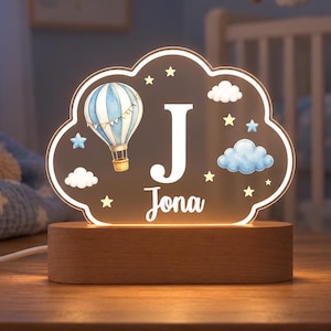 May include: A cloud-shaped LED night light with a wooden base, featuring a blue and white hot air balloon illustration, the letter J, and the name Jona in white text, surrounded by small blue and yellow stars and fluffy white and blue clouds.