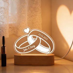 May include: A glowing LED night light featuring two intertwined wedding rings with a heart above. The rings are engraved with "John & Emily" and the date "12.11.2022". The light sits on a wooden base and casts a heart-shaped shadow.