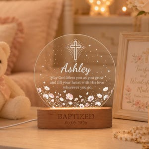 Può includere: Una luce notturna circolare in acrilico con incisioni di una croce, stelle e fiori con il testo "Ashley, May God bless you as you grow and fill your heart with His love wherever you go." La base in legno reca l'incisione "BAPTIZED 06.05.2026."