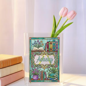 May include: A clear acrylic vase with a stained-glass design featuring a bookshelf and the name "Ashely". Two pink tulips are in the vase. Several books are stacked on the left. The stained-glass design has a teal, green, and purple color scheme.