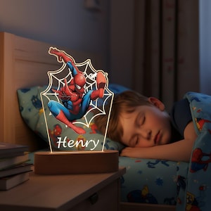 Op de afbeelding: Een Spiderman-nachtlamp met de naam "Henry" verlicht op de basis. De lamp heeft een kleurrijke Spiderman-afbeelding in een webdesign, geplaatst op een houten voet. Op de achtergrond slaapt een kind vredig.
