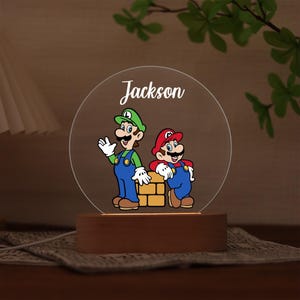 May include: A personalised night light featuring Mario and Luigi, with the name "Jackson" in white script. The lamp has a wooden base and a clear, circular acrylic panel with the cartoon characters.