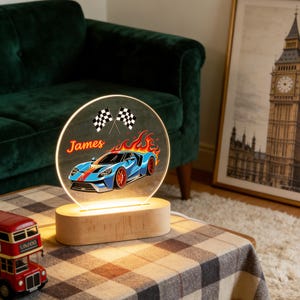 Puede incluir: Una luz de noche redonda e iluminada con un dise&ntilde;o de coche de carreras azul, llamas, banderas a cuadros y el nombre "James". La luz se encuentra sobre una base de madera y est&aacute; alimentada por un cable blanco. El tema es las carreras.