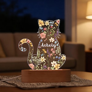 May include: A decorative cat-shaped night light with a floral design. The clear acrylic cat silhouette is filled with colorful flowers and the name "Ashely". It sits on a rectangular wooden base, illuminated by a warm glow.