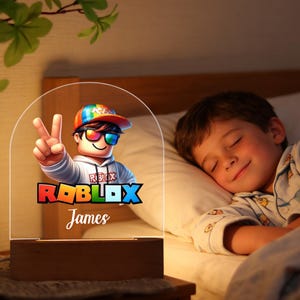 Może przedstawiać: Nocna lampka z motywem Roblox z postacią z kreskówki w okularach przeciwsłonecznych i tęczowym kapeluszu, pokazującą znak pokoju. Logo Roblox i imię "James" znajdują się poniżej. Światło stoi na drewnianej podstawie.