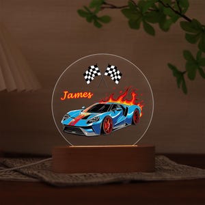 Puede incluir: Ilustraci&oacute;n de un coche deportivo azul con detalles rojos y llamas, sobre un panel acr&iacute;lico circular transparente. El nombre "James" est&aacute; escrito en naranja. Dos banderas a cuadros en la parte superior. El panel se asienta sobre una base de madera.