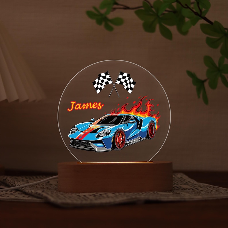 Puede incluir: Ilustraci&oacute;n de un coche deportivo azul con detalles rojos y llamas, sobre un panel acr&iacute;lico circular transparente. El nombre "James" est&aacute; escrito en naranja. Dos banderas a cuadros en la parte superior. El panel se asienta sobre una base de madera.