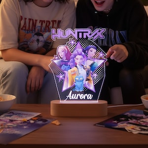 Op de afbeelding: Decoratieve LED-nachtlamp met de tekst "HUNTRAX" en de naam "Aurora". Het licht heeft een stervormig ontwerp met illustraties van drie figuren en een blauwe kat. De basis is van hout.