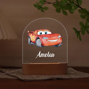 Könnte beinhalten: Ein dekoratives Nachtlicht mit einem roten Cartoon-Rennwagen mit der Nummer 95. Das Auto hat blaue Augen und ein lächelndes Gesicht. Der Name "Amelia" steht in Schreibschrift darunter. Das Licht steht auf einem Holzsockel.