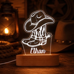 Peut inclure: Une veilleuse en acrylique illuminée présentant un graphisme en lignes blanches d'un chapeau de cowboy posé sur une paire de bottes de cowboy avec éperons. Le prénom Ethan est gravé en écriture cursive sous les bottes, sur un socle en bois.