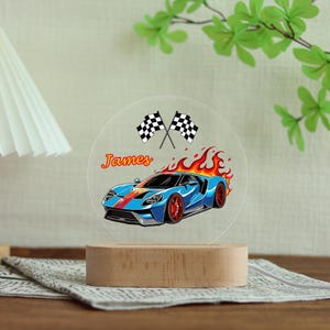 Puede incluir: Una ilustraci&oacute;n de un coche deportivo azul con llamas rojas y naranjas, el nombre "James" y banderas a cuadros se muestra en una luz nocturna redonda de acr&iacute;lico transparente. La luz se encuentra sobre una base de madera.