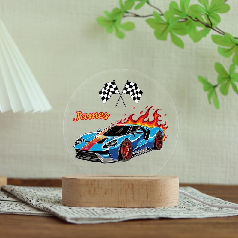 Puede incluir: Una ilustraci&oacute;n de un coche deportivo azul con llamas rojas y naranjas, el nombre "James" y banderas a cuadros se muestra en una luz nocturna redonda de acr&iacute;lico transparente. La luz se encuentra sobre una base de madera.
