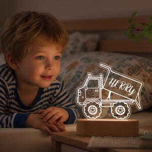 May include: A white LED truck-shaped night light with the name "HERRY" on the truck bed. The light is on a wooden base. A young child in a striped top is looking at the light, which is on a bedside table.