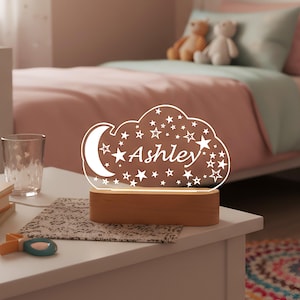 May include: A night light shaped like a cloud, with the name "Ashley" written in script. The cloud design includes a crescent moon and stars. The base is made of wood, and the light is on a white nightstand.