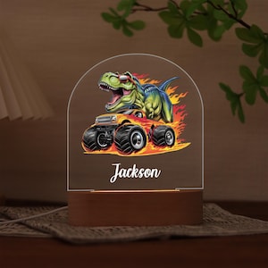 May include: A personalised night light with a cartoon dinosaur in sunglasses on a monster truck, with flames and the name "Jackson". It has a wooden base and the light is in a clear acrylic shape.
